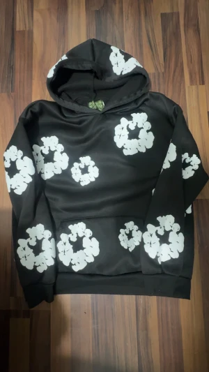 Denim Tears - Svart hoodie från Denim Tears med stora vita blommor tryckta över hela tröjan. Tröjan har huva och en klassisk känguruficka framtill. Materialet känns mjukt och skönt, perfekt för chill dagar. Snygg och unik design som sticker ut.          