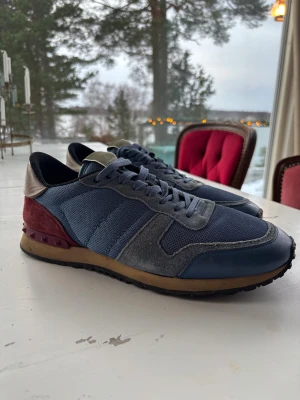 Valentino Rockrunner sneakers blå/röd - Snygga Valentino Rockrunner sneakers i blå mesh och grå mocka med röda mockadetaljer och beige skinn vid hälen. Sulan är brun med svarta nitar baktill och svart yttersula. Klassisk rund tå och snörning framtill. Perfekt för dig som gillar exklusiva sneakers med coola detaljer.