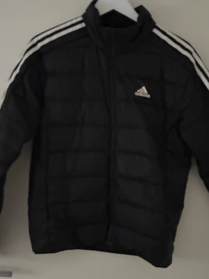Svart pufferjacka från Adidas - Svart pufferjacka från Adidas med klassiska vita ränder längs axlarna och ärmarna. Jackan har dragkedja framtill, hög krage och Adidas-logga på bröstet. Perfekt för kyliga dagar och har en sportig vibe.