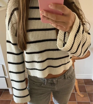 Vit och svart randig stickad tröja - Säljer en vit stickad tröja med svarta horisontella ränder. Tröjan har en croppad passform, breda långa ärmar och rund hals. Ribbstickad struktur ger en cool och avslappnad vibe. Perfekt för dig som gillar enkel men snygg stil.