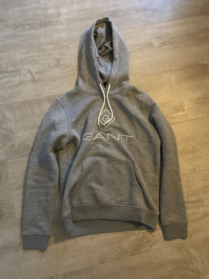 Grå hoodie från GANT med logga - Snygg grå hoodie från GANT med vit broderad logga på bröstet och vita snören med metalländar. Tröjan har huva, känguruficka och ribbade muddar vid ärmslut och nederkant. Perfekt för chill dagar och streetstyle.