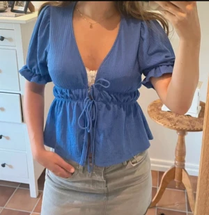 Blå omlottblus från Gina Tricot - Säljer en blå blus från Gina Tricot med omlott-design och snörning i midjan. Blusen har puffiga korta ärmar med resår och en djup v-ringning. Materialet är lätt och strukturerat, perfekt för vår och sommar. Supersnygg till jeans eller kjol!