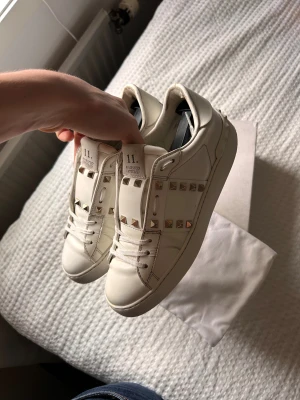 Valentino Rockstud vita sneakers - Säljer ett par vita Valentino Rockstud sneakers i skinn med ikoniska nitar längs sidorna och bak på sulan. Skorna har rund tå, platt sula och vita snören. Perfekt för dig som vill ha en clean men edgy look med exklusiv känsla.