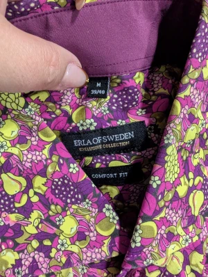 Blommig skjorta från Erla of Sweden stl M - Färgstark skjorta från Erla of Sweden i comfort fit med blommigt mönster i lila, rosa och gulgrönt. Skjortan har klassisk krage, långa ärmar och knäppning framtill. Perfekt för dig som vill sticka ut med en unik och lekfull stil.