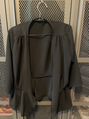 Svart cardigan jacka från Boohoo - Svart cardigan jacka från Boohoo med volangkant nertill och öppen framtill. Jackan har trekvartsärmar och är tillverkad i ett lätt, strukturerat tyg som ger en snygg och avslappnad look. Perfekt för att slänga över en topp eller klänning.