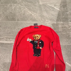 Röd Polo Bear tröja från Ralph Lauren - Riktigt snygg röd stickad tröja från Polo Ralph Lauren med ikoniska Polo Bear på bröstet, klädd i kostym och rutig halsduk. Rund hals och RL-broderi vid nederkant. Materialmix med merinoull, nylon, akryl och polyester. Perfekt statement piece för vintern och julen.