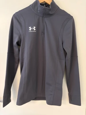 Grå half zip träningströja Under Armour - Sportig grå långärmad träningströja från Under Armour med half zip och hög krage. Snyggt logotryck på bröstet och smidig passform som funkar perfekt till gymmet eller löprundan. Materialet är mjukt och stretchigt syntet för maximal komfort.
