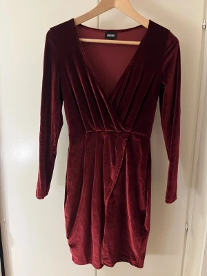 Vinröd omlottklänning från Bubbleroom - Supersnygg vinröd kortklänning från Bubbleroom i mjuk sammet. Klänningen har omlott-design med v-ringning och långa ärmar. Perfekt för dig som gillar lyxig känsla och elegant stil. Passformen är normal och tyget har ett fint skimmer.