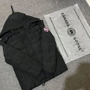 Svart dunjacka från Canada Goose - Säljer en svart dunjacka från Canada Goose med huva och klassisk logga på ärmen. Jackan har långa ärmar, knappar framtill och är perfekt för kalla dagar. Snygg och stilren design som håller dig varm hela vintern.