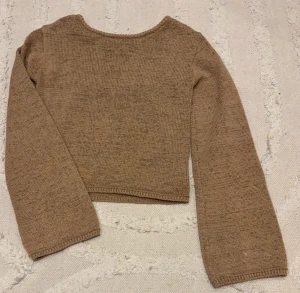 Grön brun stickad tröja - Säljer en grön brun  stickad tröja ifrån Indiska med långärmad design och öppen rygg. Tröjan har en snygg omlott-detalj bak som ger en cool och trendig look. Perfekt för dig som vill ha något som sticker ut lite extra.