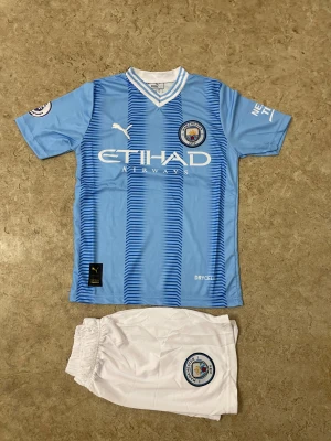 Manchester City Haaland #9 matchtröja - Manchester Citys officiella matchtröja med Haaland och nummer 9 på ryggen. Tröjan är ljusblå med vita detaljer, korta ärmar och randigt mönster framtill. Materialet är lätt och andas, perfekt för fotboll. Ingår vita shorts med klubbmärke.