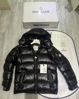 Svart Moncler pufferjacka med huva - Svart pufferjacka från Moncler med glansig finish, huva och klassisk logga på ärmen. Jackan har dragkedja framtill, två fickor med dragkedja och quiltad design. Perfekt för kalla dagar och riktigt snygg streetstil.