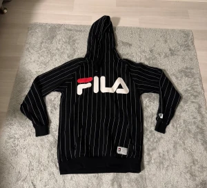 Svart randig hoodie från FILA - Svart hoodie från FILA med vita tunna ränder och stor logga i vitt och rött på bröstet. Tröjan har huva med snörning, långärmade ribbade muddar och en praktisk magficka. Perfekt för chill eller streetwear-stil.