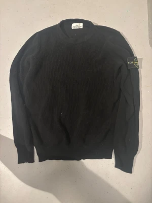 Svart stickad tröja Stone Island  - Svart stickad tröja från Stone Island i storlek S. Klassisk rund hals och ribbad struktur. Ikonisk Stone Island patch på vänster ärm. Perfekt för dig som gillar clean streetwear med premiumkänsla.