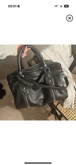 Svart handväska från Zadig & Voltaire - Cool svart handväska i skinn från Zadig & Voltaire med silverfärgade dragkedjor och nitar längs kanterna. Väskan har både handtag och axelrem med broderad logga. Perfekt storlek för att få plats med allt viktigt och har en edgy vibe. Den är i den stora modellen