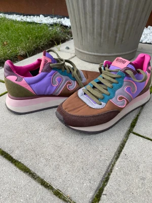Färgglada Wushu Ruyi sneakers - Säljer ett par unika sneakers från Wushu Ruyi med cool färgkombo i lila, rosa, brun, grön och blå. Skorna har snörning, platt sula och rund tå. Materialmix med mocka, syntet och textil. Snygga detaljer med vågiga mönster på sidorna och kontrastfärgad innersula.