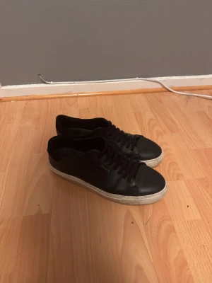 Svarta sneakers från Axel Arigato - Stilrena svarta sneakers från Axel Arigato i skinn med vit platt sula och diskret guldfärgad logga på sidan. Klassisk rund tå och snörning framtill. Perfekta för dig som gillar minimalistisk och clean stil.