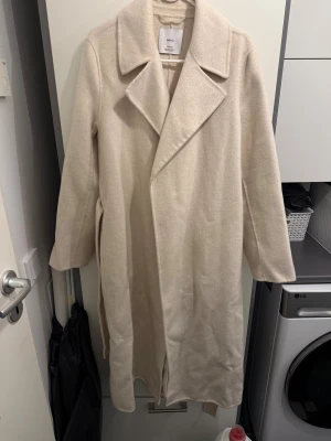 Beige kappa från MNG med bälte - Stilren beige kappa från MNG med klassisk krage och bälte i midjan. Lång modell med raka ärmar och mjukt ullmaterial. Perfekt för dig som gillar en clean och tidlös look.
