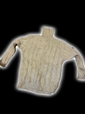 Beige polotröja från Paul & Shark - Säljer en beige polotröja från Paul & Shark med grov kabelstickning och ribbade muddar. Tröjan har hög polokrage och lång ärm, perfekt för kalla dagar. Materialet är mjuk ull och den har en snygg logotyp på ärmen.