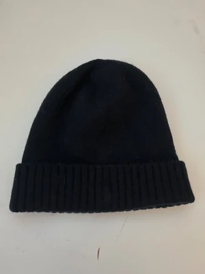 Svart ullmössa från Polo Ralph Lauren - Klassisk svart mössa från Polo Ralph Lauren i 100% ull. Mössan har en ribbstickad uppvikt kant och enkel, stilren design som passar till allt. Perfekt för kalla dagar när du vill hålla stilen enkel men snygg. För de mindre, töjs ut ganska mycket 