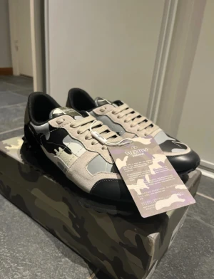 Valentino Garavani Rockrunner sneakers - Valentino Garavani Rockrunner sneakers med grå, beige och svarta camouflagemönster. Skorna har snörning, platt sula med nitar och rund tå. Materialmix av mocka och skinn ger en lyxig känsla. Kommer med originalkartong, extra skosnören och dustbag.  Ny pris 7000kr