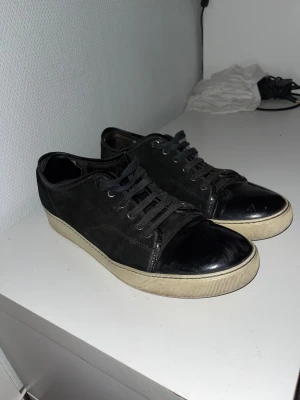 Svarta sneakers från Lanvin med lackad tå - Säljer ett par svarta Lanvin sneakers med snygg lackad tå och vit platt sula. Skorna har snörning och är tillverkade i mocka med detaljer i skinn. Klassisk rund tå och stilren design som passar till många outfits. Dustbag från Lanvin medföljer. Var inte rädda att höra av er om ni undrar något. Pris kan diskuteras 