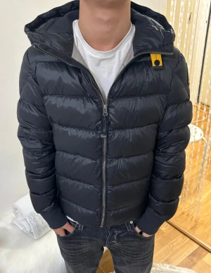 Svart dunjacka Parajumpers Sheen Y-M - Säljer en svart dunjacka från Parajumpers, modell Sheen. Jackan har en glansig finish, huva, ribbade muddar och dragkedja framtill. Klassisk PJS-patch på ärmen och gul detalj vid halsen. Insidan är ljusgrå och jackan är riktigt varm och stilren. Pris kan diskuteras!