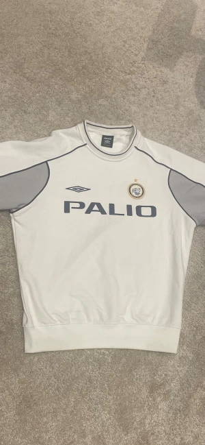 Vit sweatshirt Palace x Umbro - Vit och grå långärmad sweatshirt från Palace x Umbro med tryck på både fram- och baksida. Tröjan har mörkblå detaljer, logga och emblem på bröstet samt kontrastpaneler på ärmarna. Perfekt för dig som gillar streetwear och sportig stil. Tveka inte på att skriva vid frågor.