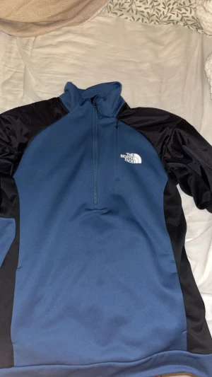 Blå half zip hoodie från The North Face - Snygg blå hoodie från The North Face med svarta detaljer på axlar och sidor. Tröjan har half zip framtill, långärmade muddar och logga på bröstet. Perfekt för träning eller chill, tillverkad i mjuk polyester som känns skön mot huden. den är köpt för 2200