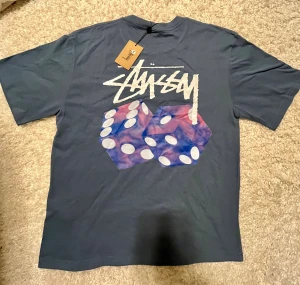 Blå Stüssy x NIKE tröja  - Helt nya snygg blå t-shirt från Stüssy med stort vitt logotryck och två färgglada tärningar på ryggen. Framtill finns en mindre Stüssy-logga. Tillverkad i 100% bomull med klassisk passform och korta ärmar. Perfekt för dig som gillar streetwear och coola prints.