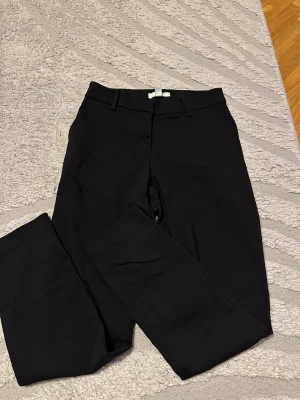 Svarta kostymbyxor med dragkedja - Snygga svarta kostymbyxor med klassisk rak passform och midwaist. Byxorna har bälteshällor, framfickor och två bakfickor. Stängs med dragkedja och två hyskor. Perfekta för en clean och stilren look.