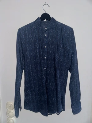 Mönstrad blå skjorta från 2BLIND2C - Snygg blå skjorta från 2BLIND2C i slim fit med ett vågigt mönster i olika blå nyanser. Skjortan har långa ärmar, klassisk krage och vita knappar framtill. Perfekt för dig som vill sticka ut lite extra med en cool design.