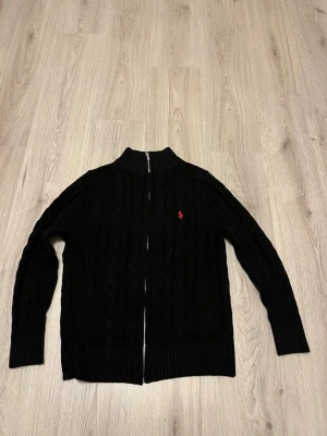 Svart kabelstickad kofta Polo Ralph Lauren - Svart kabelstickad kofta från Polo Ralph Lauren med röd broderad logga på bröstet. Koftan har ribbad krage, muddar och nederkant samt hel dragkedja framtill. Materialet är mjuk bomull och passformen är normal. Perfekt för dig som gillar klassisk stil med en twist.