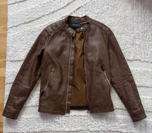 Skinnjacka - Snygg brun skinnjacka från Pull&Bear med raka linjer och coola detaljer på axlarna. Jackan har två snedställda fickor med dragkedja och en stilren ståkrage med tryckknapp. Perfekt för dig som vill ha en klassisk bikerstil.