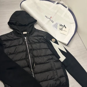 Svart pufferjacka, Moncler - Svart pufferjacka från Moncler med huva och dragkedja framtill. Jackan har stickade ärmar med vit zigzag-mönster och Moncler-logga på ena ärmen. Kroppen är quiltad och fylld med dun, medan ärmarna är i mjuk ull. Snygg med coola detaljer. Jag har använd den ett tag ca 7 måndagar men inga märken syns att den är använd. Det enda är att NFC scan funkar inte längre då den gick sönder i tvätten annars en väldigt fin puffer jacka/kofta. Allt ni ser på bilder följer med. Kom DM för mer bilder :)