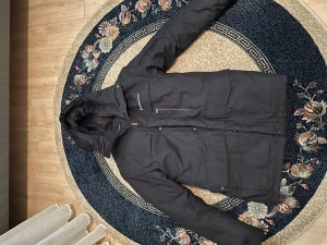 Svart dunjacka Peak Performance - Peak Performance svart dunjacka med huva och quiltad insida. Jackan har flera stora fickor, dragkedja och tryckknappar framtill. Insidan av huvan är fodrad med mjuk pälsimitation för extra komfort. Perfekt för kalla vinterdagar.