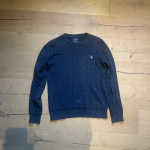 Mörkblå kabelstickad tröja GANT - Snygg mörkblå kabelstickad tröja från GANT med rund hals och diskret logga broderad på bröstet. Tillverkad i mjuk premium bomull och har ribbade muddar vid ärmslut och nederkant. Perfekt för dig som gillar klassisk stil.