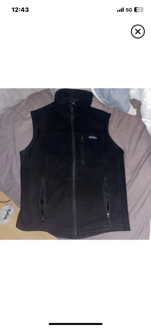 Svart fleeceväst från Patagonia Synchilla - Svart fleeceväst från Patagonia Synchilla i storlek S. Västen har hög krage, tre fickor med dragkedja och en stilren look. Perfekt att slänga på över en hoodie eller t-shirt när det är lite kyligt ute. Mjuk och skön fleece som håller värmen.