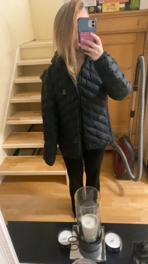Svart dunjacka från Haglöfs - Svart dunjacka från Haglöfs med snygg quiltad design och dragkedja framtill. Jackan har huva och lång ärm, perfekt för kalla dagar. Klassisk och stilren look med diskret logga på bröstet. Passar dig som vill ha en varm och lätt jacka med skön passform. Finns ett hål vid ena ärmen, se bild 5. 