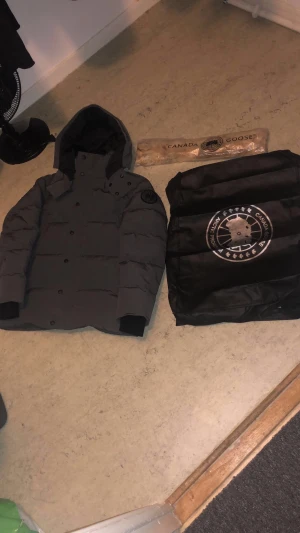 Äkta grå canada goose jacka  - Säljer en mörkgrå dunjacka från Canada Goose med huva och svart logotyp på ärmen. Jackan har raka linjer, knappar framtill och ribbade muddar. Perfekt för kalla dagar och riktigt snygg streetstil.