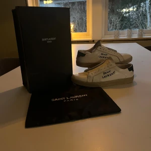 Saint Laurent vita sneakers i skinn - Säljer ett par vita Saint Laurent sneakers i skinn med svart häl och handskriven logga på sidan. Klassisk låg modell med rund tå och platt sula. Skorna kommer med originalkartong och dustbag. Perfekta för dig som gillar stilrena och exklusiva sneakers.
