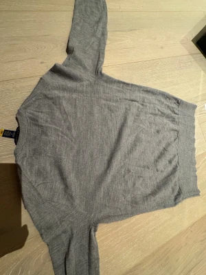 Grå stickad tröja från Polo Ralph Lauren - Snygg grå stickad tröja från Polo Ralph Lauren med klassisk rund hals och broderad logga på bröstet. Tröjan har långärmad passform och ribbade muddar vid ärmslut och nederkant. Perfekt för dig som gillar stilrena och tidlösa plagg.