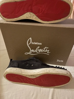 Christian Louboutin svarta sneakers med nitar - Grå sneakers från Christian Louboutin med ikoniska röda sulor och grå nitar på tån. Skorna har snörning och är tillverkade i mocka med platt sula. Snygg och edgy design som sticker ut med sina detaljer och exklusiva känsla. Några spikar fattas men går att hitta på nätet. Pris kan diskuteras. Tveka inte att fråga…