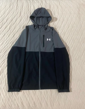 Grå och svart vindjacka Under Armour - Snygg vindjacka från Under Armour i grått och svart med huva och hel dragkedja. Jackan har en bröstficka med dragkedja och logga på framsidan. Elastiska muddar med Under Armour-tryck. Perfekt för träning eller chill ute.