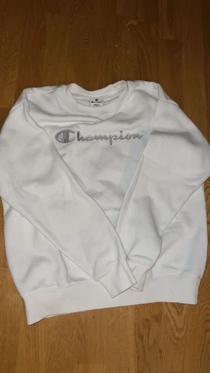 Vit sweatshirt från Champion, S - Vit sweatshirt från Champion i storlek small. Tröjan har ett broderat Champion-logo i grått och vitt framtill, rund hals och ribbade muddar vid ärmslut och nederkant. Mjuk insida i bomull och polyester, perfekt för chill dagar.