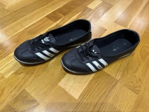 Svarta Adidas ballerina sneakers - Säljer ett par svarta Adidas ballerina sneakers med klassiska vita ränder på sidorna och snörning framtill. Skorna har rund tå och platt sula, tillverkade i syntetmaterial med perforerade detaljer för extra stil. Perfekt för dig som gillar sportig och clean look.