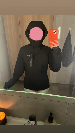 Svart skidjacka från Helly Hansen - Svart skidjacka från Helly Hansen med huva. Jackan har en snygg, minimalistisk design med HH-logga på bröstet och reflekterande detaljer.  Endast använd några ggr förra vintern så den är nästan som helt ny. Den har några få fläckar som knappt syns och som jag är ganska säker på går att tvätta bort. Nypris 2999. Sååå snygg!💕