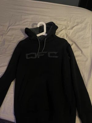Svart hoodie med QFC-tryck Orsa fiske center strl xs - Svart hoodie från QFC med stort tryck på bröstet. Tröjan har huva med snörning och en klassisk magficka framtill. Långa ärmar och ribbade muddar ger en skön och avslappnad look. Perfekt för chill dagar eller när du vill ha något bekvämt.