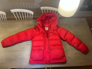 Röd dunjacka från Fjällräven - Fin röd dunjacka från Fjällräven med stor huva, gula band och två stora fickor framtill. Klassisk badge på bröstet och snygga detaljer i gult. Perfekt för kalla dagar när du vill sticka ut.