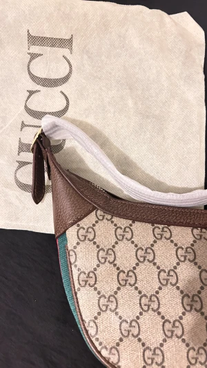 Gucci axelväska med GG-mönster - Snygg axelväska från Gucci med klassiskt GG-mönster i beige och brunt. Väskan har bruna skinn-detaljer, guldfärgad GG-logga och dragkedja. Sidan har röd och grön rand. Perfekt för dig som vill ha en ikonisk designeraccessoar med lyxig känsla.